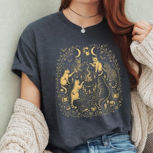 Op de afbeelding: Een donkergrijs T-shirt met korte mouwen en een goudkleurige grafische print van vijf katten die zich rond een centrale ketel verzamelen. Het ontwerp bevat maanfasen, kleine pompoenen, kaarsen en een boek met een pentagramsymbool.