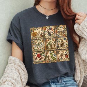 Può includere: Una t-shirt grigio antracite scuro con una griglia di nove illustrazioni quadrate in stile manoscritto medievale. Le grafiche raffigurano vari insetti e piccole creature, tra cui una lumaca, uno scarabeo, una farfalla, una libellula, una mantide, un'ape, formiche, un bruco e un ragno, circondati da bordi floreali decorati.