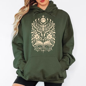 May include: An olive green hooded sweatshirt featuring a cream-colored graphic print of a moth, an open book, flowers, and celestial elements. The text "THE STORYTELLER" is printed in a serif font below the open book illustration.