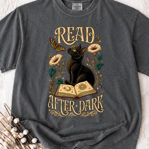 Puede incluir: Una camiseta gris carbón con un estampado gráfico de un gato negro con gafas, sentado detrás de un libro abierto con símbolos celestiales. El diseño incluye una polilla y flores, con el texto READ AFTER DARK en una fuente decorativa.