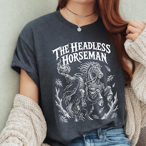 May include: A dark charcoal gray t-shirt featuring a white graphic print of the Headless Horseman riding a horse while holding a flaming pumpkin. The text THE HEADLESS HORSEMAN is printed in a stylized font above the detailed illustration.