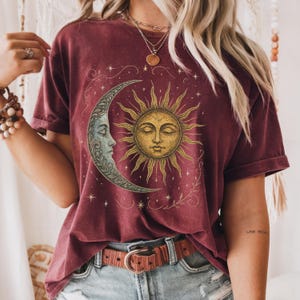 Op de afbeelding: Bordeauxrood T-shirt met een hemels ontwerp. De afbeelding bevat een maan met een gezicht en een zon met een gezicht, omringd door sterren en bloemmotieven. Het shirt heeft korte mouwen en een relaxte pasvorm.