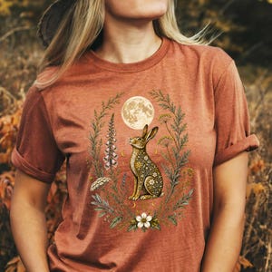 May include: A rust-colored short-sleeve t-shirt featuring a central graphic of a rabbit sitting within a circular wreath of wildflowers and foliage. Above the rabbit is a full moon, and the rabbit's body is decorated with intricate floral patterns.