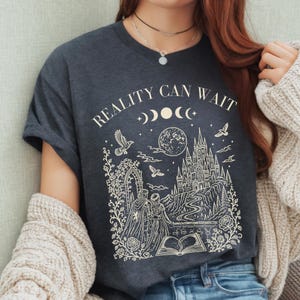 May include: A dark charcoal grey t-shirt featuring a cream-coloured graphic print. The design includes the text "REALITY CAN WAIT" above a moon phase cycle, a castle, a full moon, birds, a couple, and an open book, all framed by floral vines.