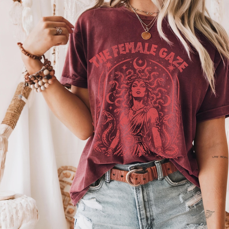 Pu&ograve; includere: T-shirt bordeaux con grafica rossa di Medusa e la scritta "THE FEMALE GAZE". Il design include serpenti, stelle e una luna crescente. La maglietta &egrave; abbinata a pantaloncini di jeans e una cintura marrone.