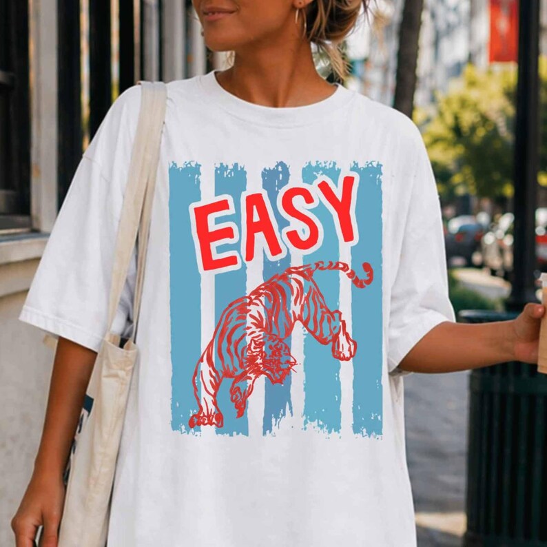 May include: A white short-sleeved t-shirt featuring a graphic print with the word EASY in bold red letters above a red tiger illustration. The design is set against a background of vertical light blue stripes on the front of the garment.