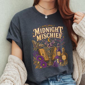 Może przedstawiać: Ciemnoszary t-shirt z nadrukiem graficznym z napisem MIDNIGHT MISCHIEF złotymi literami. Wzór zawiera centralnie umieszczony kielich, otwartą księgę, dwie szklane butelki, ćmę, fioletowe kwiaty, grzyby oraz małe nietoperze w tle.