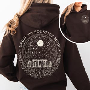 Puede incluir: Sudadera con capucha marrón oscuro con un gráfico blanco en la espalda. El gráfico incluye el texto "UNDER THE SOLSTICE MOON" y un diseño de la luna, las estrellas y Stonehenge. La sudadera tiene capucha y bolsillo delantero.