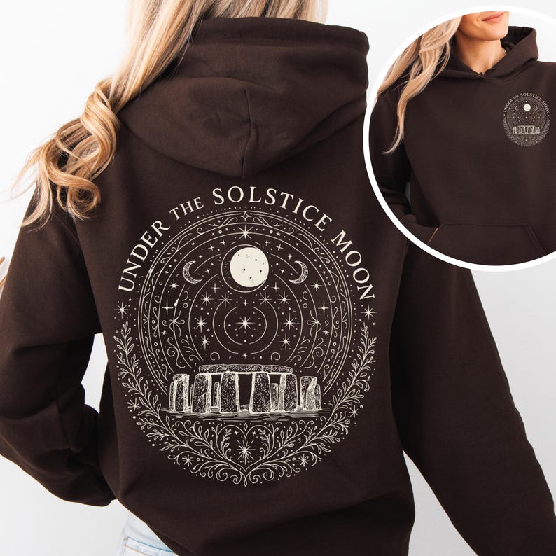 May include: A dark brown hoodie with a white graphic on the back featuring the text "UNDER THE SOLSTICE MOON" around a design of the moon, stars, and Stonehenge. The hoodie has a hood and a front pocket.