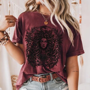 Op de afbeelding: Bordeauxrood t-shirt met een zwarte afbeelding van Medusa, met slangen als haar, omgeven door een bloemen- en maansikkelontwerp. Het t-shirt heeft korte mouwen en een relaxte pasvorm.