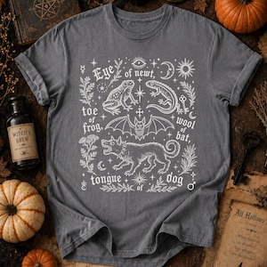 May include: A charcoal gray t-shirt featuring a white graphic print of a frog, a newt, a bat, and a dog, surrounded by botanical elements, celestial symbols, and the text: Eye of newt, toe of frog, wool of bat, tongue of dog.