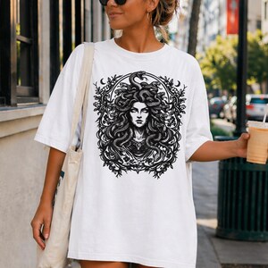 Op de afbeelding: Wit T-shirt met een zwart-wit afbeelding van Medusa, met slangen als haar, omringd door een bloemenkrans. De afbeelding is gecentreerd. Het shirt heeft korte mouwen en een ronde hals.