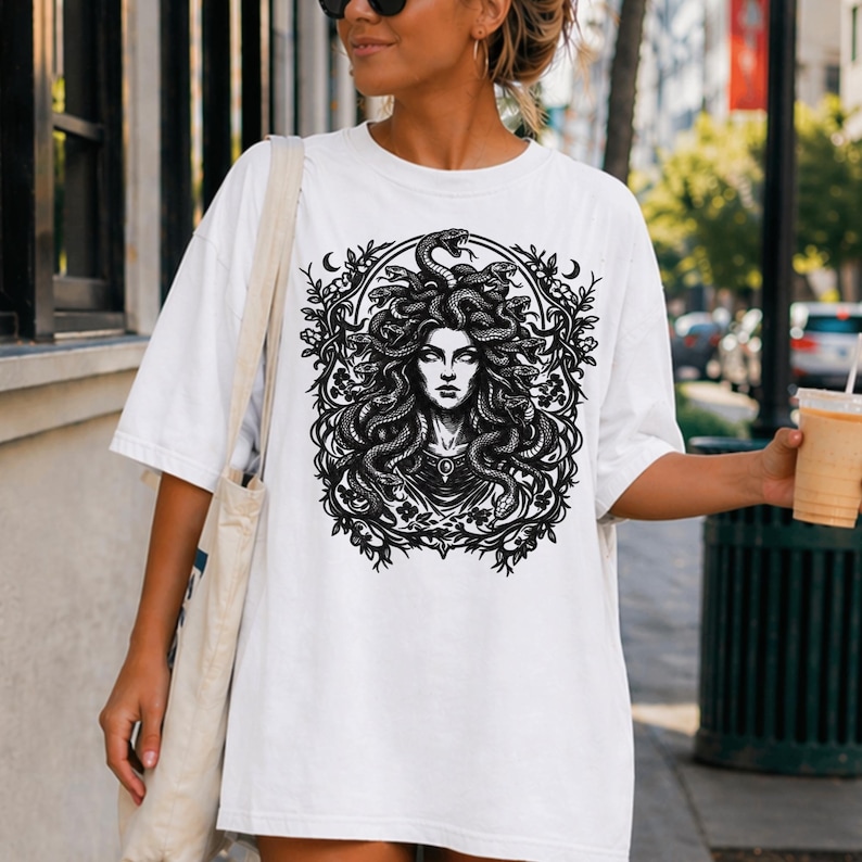 Op de afbeelding: Wit T-shirt met een zwart-wit afbeelding van Medusa, met slangen als haar, omringd door een bloemenkrans. De afbeelding is gecentreerd. Het shirt heeft korte mouwen en een ronde hals.