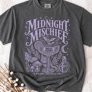 May include: A charcoal gray t-shirt featuring a light purple graphic print with the text "MIDNIGHT MISCHIEF" and "WITCHING HOUR SOCIAL CLUB". The design includes a stemmed glass, a cat, a moth, mushrooms, bats, and a crescent moon.