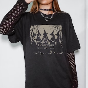 May include: A black short-sleeved t-shirt featuring a graphic print of five figures in pointed hats standing in a circle. The text reads The Real Housewives of Pendle Hill. The shirt is worn over a black long-sleeved mesh top with a chain necklace.