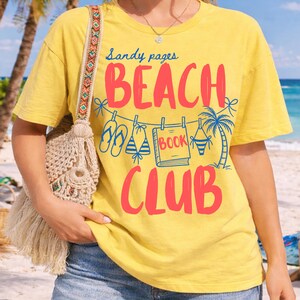 May include: Yellow t-shirt with the words "Sandy pages BEACH BOOK CLUB" in red and blue. The design includes illustrations of flip-flops, a bikini, a book, and a palm tree. The shirt is a casual, beach-themed design.