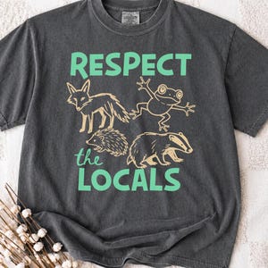 Può includere: T-shirt grigio scuro con le parole "RESPECT the LOCALS" in verde. La maglietta presenta disegni a tratto di una volpe, una rana, un riccio e un tasso. La maglietta è realizzata in un materiale morbido e confortevole.