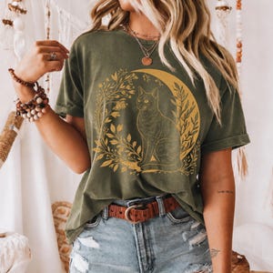 Op de afbeelding: Olijfgroen T-shirt met een gouden illustratie van een kat in een maan en omringd door bladeren. Het shirt heeft korte mouwen en een ronde hals. De persoon draagt een bruine riem en een spijkerbroek.