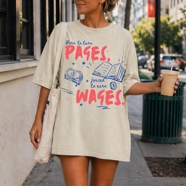 May include: Cream-colored t-shirt with the text "Born to turn PAGES, forced to earn WAGES." The design includes illustrations of a stack of money and an open book, all in pink and blue. The shirt has short sleeves.