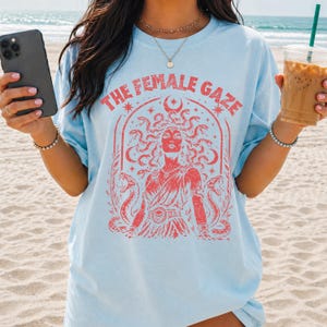Pu&ograve; includere: T-shirt azzurra con la scritta "THE FEMALE GAZE" in rosso. La grafica presenta un'illustrazione rossa di una donna con serpenti al posto dei capelli, circondata da serpenti e stelle. La maglietta &egrave; in stile casual e unisex.