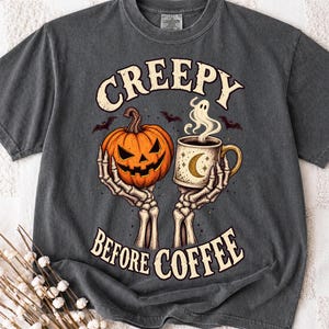 May include: A charcoal grey t-shirt featuring a graphic of two skeleton hands holding an orange jack-o'-lantern and a coffee mug with a crescent moon. The text reads CREEPY BEFORE COFFEE, surrounded by small bats and a ghost rising from the mug.