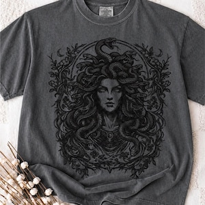 Op de afbeelding: Donkergrijs T-shirt met een zwart Medusa-ontwerp, omringd door een bloemenpatroon. Het ontwerp toont Medusa met slangen als haar. Het T-shirt heeft een ronde hals en korte mouwen.