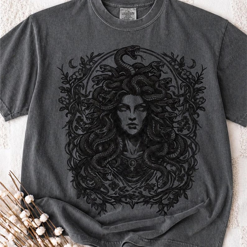 Op de afbeelding: Donkergrijs T-shirt met een zwart Medusa-ontwerp, omringd door een bloemenpatroon. Het ontwerp toont Medusa met slangen als haar. Het T-shirt heeft een ronde hals en korte mouwen.