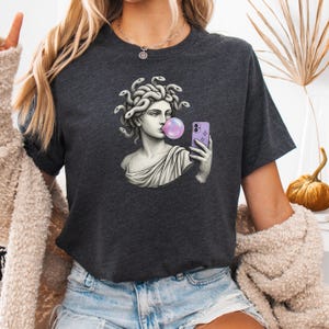 May include: Dark grey t-shirt with a graphic of Medusa blowing a bubblegum bubble while taking a selfie with a purple phone. The Medusa graphic is in a classical statue style.