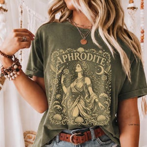 May include: An olive green t-shirt featuring a cream-coloured graphic print of a figure with long flowing hair, surrounded by ornate floral patterns, seashells, birds, and a crescent moon, with the text APHRODITE arched above the central figure.