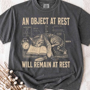 Puede incluir: Una camiseta gris carbón con un gráfico de un gato atigrado durmiendo sobre un libro abierto. El diseño incluye el texto AN OBJECT AT REST WILL REMAIN AT REST, un despertador vintage, una pluma de escribir y diagramas de física con etiquetas.