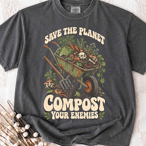 May include: A charcoal gray t-shirt featuring a graphic of a wheelbarrow filled with garden waste, a skull, and mushrooms. The text reads "SAVE THE PLANET" at the top and "COMPOST YOUR ENEMIES" at the bottom in a bold, cream-colored retro font.