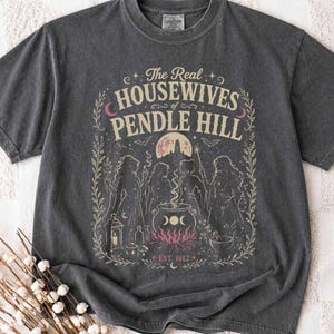 May include: A charcoal gray short-sleeve t-shirt featuring a graphic print with the text The Real HOUSEWIVES of PENDLE HILL EST. 1612. The design includes silhouettes of four figures around a cauldron, a full moon, bats, and botanical borders.