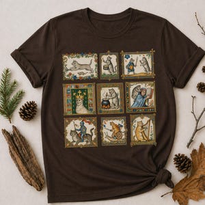 Op de afbeelding: Een donkerbruin t-shirt met een raster van negen rechthoekige illustraties met antropomorfe katten in middeleeuwse sc&egrave;nes, waaronder een kat met een kroon, een kat die harp speelt, een kat op een slak en een kat die in een pot kookt.
