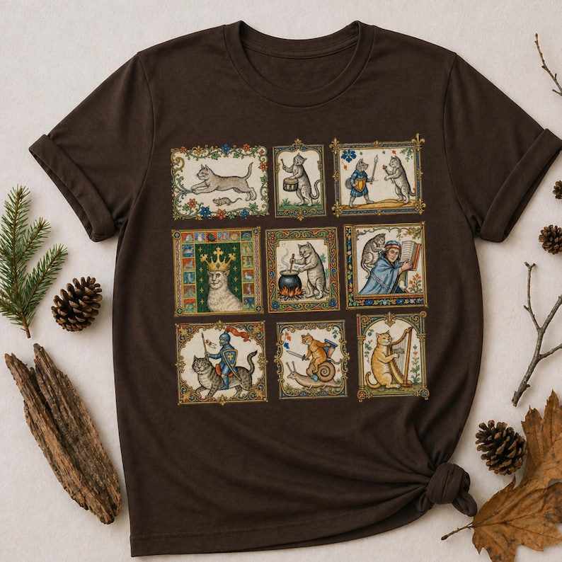 Op de afbeelding: Een donkerbruin t-shirt met een raster van negen rechthoekige illustraties met antropomorfe katten in middeleeuwse sc&egrave;nes, waaronder een kat met een kroon, een kat die harp speelt, een kat op een slak en een kat die in een pot kookt.