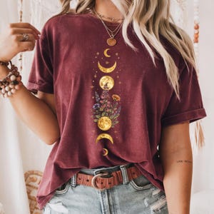 Op de afbeelding: Bordeauxkleurig T-shirt met een hemels ontwerp van maanfasen en bloemelementen. De afbeelding staat in het midden van de voorkant. Het shirt heeft korte mouwen en een relaxte pasvorm, gecombineerd met een bruine riem en een spijkerbroek.