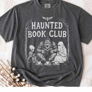 Può includere: Una t-shirt a maniche corte grigio antracite con una stampa grafica bianca raffigurante un alieno, un sasquatch e un fantasma seduti insieme con libri e tazze da tè. Il testo HAUNTED BOOK CLUB è stampato sopra le figure.
