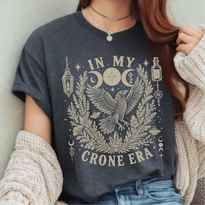 May include: A dark grey t-shirt featuring a cream-coloured graphic of a raven with wings spread, surrounded by botanical elements, a lantern, a bottle, and moon phases. The text "IN MY CRONE ERA" is printed in a bold, serif font above and below the raven.