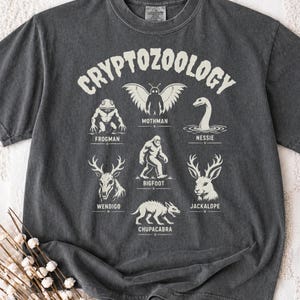 May include: A charcoal grey t-shirt featuring a cream-coloured graphic with the text CRYPTOZOOLOGY at the top. The design displays illustrations of Mothman, Frogman, Nessie, Bigfoot, Wendigo, Jackalope, and Chupacabra with their names printed below.