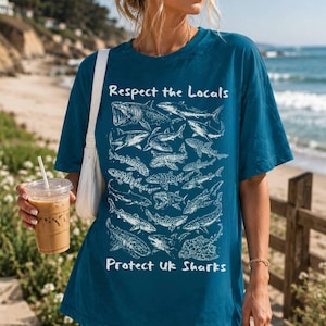 Könnte beinhalten: Türkisfarbenes T-Shirt mit weißen Hai-Illustrationen. Der Text "Respect the Locals" und "Protect UK Sharks" ist weiß gedruckt. Das Shirt hat eine lockere Passform.