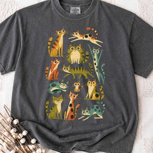 Puede incluir: Una camiseta gris carbón de manga corta con un estampado gráfico de once gatos estilizados en varias poses y colores, incluyendo naranja, verde y azul cerceta. Los gatos tienen ojos grandes y patrones distintos, dispuestos en un grupo.