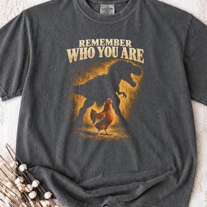 May include: A charcoal gray short-sleeve t-shirt featuring a graphic print of a brown chicken standing in front of a large, dark silhouette of a Tyrannosaurus Rex. The text "REMEMBER WHO YOU ARE" is printed in a bold, light-colored font above.