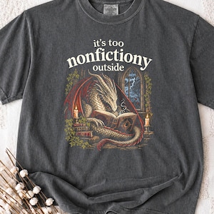 Pode incluir: Uma t-shirt cinzento-carvão com uma estampa gráfica de um dragão a ler um livro à luz de velas. O texto acima do dragão diz "it's too nonfiction outside". A ilustração inclui pilhas de livros, ramos de hera e uma vista de uma janela.