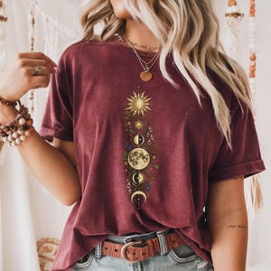 Pode incluir: Uma t-shirt de manga curta cor de vinho com uma impressão gráfica vertical de um sol, várias fases da lua e elementos florais coloridos. O design está centrado no peito, exibindo corpos celestes rodeados por folhas verdes e pequenas flores.