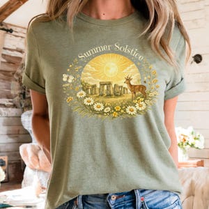 May include: Olive green t-shirt featuring a circular graphic with the text "Summer Solstice." The design includes a sun, Stonehenge, a deer, butterflies, and flowers. The shirt has short sleeves and a crew neck.