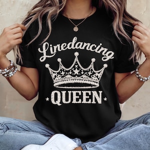 Op de afbeelding: Zwart t-shirt met de witte tekst "Linedancing QUEEN" en een kroonontwerp. De kroon is gedetailleerd met sterren en parelachtige accenten. Een casual t-shirt voor dagelijks gebruik.