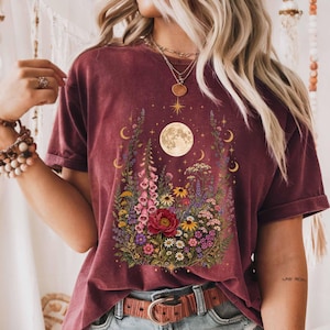 Puede incluir: Una camiseta de manga corta color granate con un estampado gráfico central que muestra una luna llena, una estrella y un vibrante arreglo de flores silvestres coloridas, incluyendo dedaleras rosas, peonías rojas, margaritas amarillas y flores moradas.