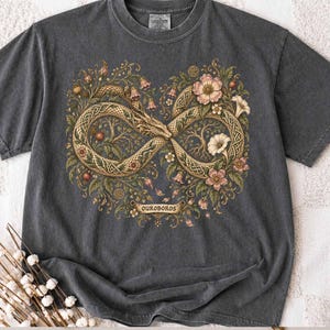 May include: A charcoal grey short-sleeved t-shirt featuring a graphic of an infinity-shaped serpent with intricate Celtic knotwork patterns, surrounded by detailed botanical illustrations of flowers and leaves, with the text OUROBOROS below.