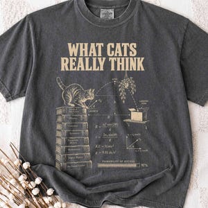 Può includere: Una t-shirt grigio antracite a maniche corte con una stampa grafica color crema. Il design mostra un gatto seduto su una pila di libri, un diagramma di traiettoria fisica e il testo WHAT CATS REALLY THINK, insieme a varie equazioni scientifiche.