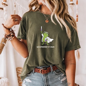 May include: An olive green short-sleeve t-shirt featuring a graphic of a green frog wearing a white lab coat and goggles, holding a test tube and beaker with purple liquid. The text below the frog reads, WE EXPERIMENT AT DAWN.