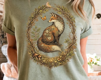 May include: Sage green t-shirt featuring a detailed illustration of a fox within a floral wreath. The fox has brown and white fur, adorned with floral patterns. A butterfly and a crescent moon are also included in the design. The shirt is a casual wear item.